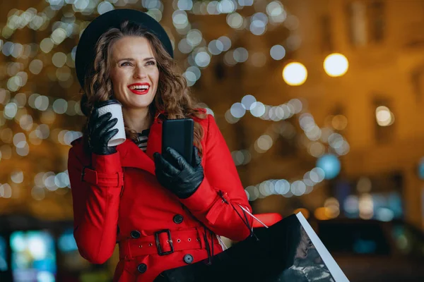 冬天的乐趣 穿着红色外套 头戴黑色贝雷帽 头戴购物袋 头戴茶杯和智能手机 晚上在城外用应用软件的现代单身女性微笑着 — 图库照片