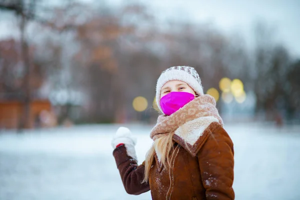 ニット帽とシープスキンコートでミトンと医療マスクを持つスタイリッシュな子供の笑顔の肖像は冬に市内公園の外で雪玉を果たしている — ストック写真