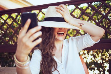 40 yaşında, beyaz gömlekli mutlu bir ev hanımı. Pansiyonun terasındaki akıllı telefondan video konferansı yapıyor..