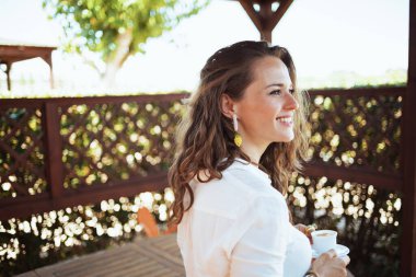 Gülümseyen modaya uygun 40 yaşındaki beyaz gömlekli ev kadını terasta bir fincan kahveyle.