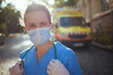Covid-19 salgını. Dışarıda ambulansın yanında steteskop ve tıbbi maske takan, üniformalı, düşünceli, modern sağlık görevlisi kadın..