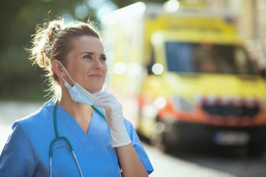 Covid-19 salgını. Steteskoplu ve sağlık maskeli modern tıp doktoru gülümsüyor. Dışarıda ambulansın yanında nefes alıyor..
