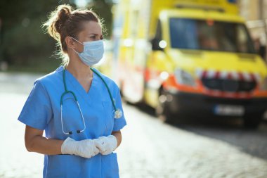 Covid-19 salgını. Steteskop ve tıbbi maskeli, ambulansın yanında üniformalı, düşünceli, modern tıp doktoru kadın..