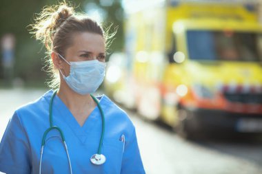 Covid-19 salgını. Steteskop ve tıbbi maske takmış modern doktor kadın ambulansın yakınındaki uzaklığa bakıyor..