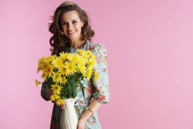 Uzun dalgalı esmer saçlı, gülümseyen stil sahibi bir kadının portresi sarı kasımpatı çiçekleri ile pembe arka planda duran vazoda..