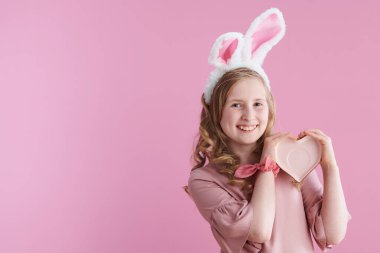 Pembe elbiseli, gülümseyen stil bir çocuğun portresi. Kalp şeklinde fincan tabağı ve pembe arka planda tavşan kulakları var..