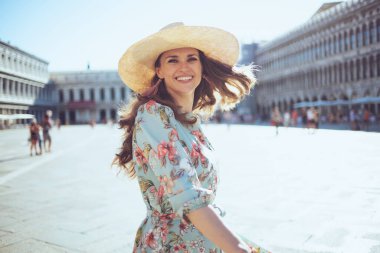 Gülümseyen, çiçek desenli elbiseli, şapkalı modern kadın Venedik, İtalya 'daki Piazza San Marco' da gezintiye çıkıyor..