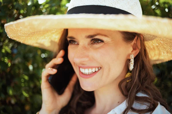 40岁穿着白衬衫 头戴帽子 在户外绿墙附近的智能手机上说话的快乐而时尚的女人 — 图库照片