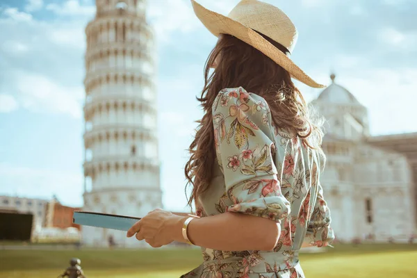 Seen from behind young traveller woman in floral dress with pizza and hat near Leaning Tower in Pisa, Italy.