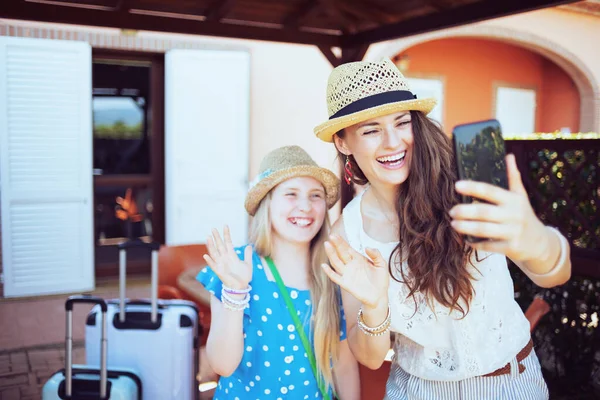 幸せなスタイリッシュなお母さんと娘で帽子でトロリーバッグを持っていますウェブカメラミーティングでゲストハウスホテルのパティオでスマートフォン — ストック写真