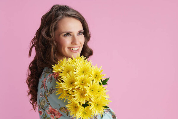 smiling modern 40 years old woman with long wavy brunette hair with yellow chrysanthemums flowers isolated on pink background.