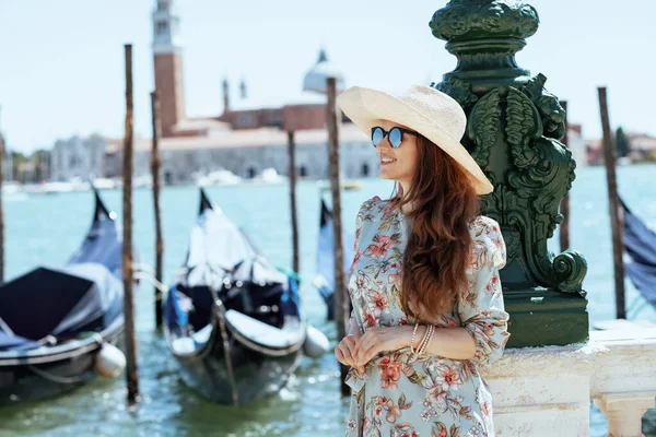 サングラスと帽子を持つ花のドレスで流行のソロ旅行者の女性を笑顔ヴェネツィア イタリアの堤防の上を歩くツアーを持っている — ストック写真