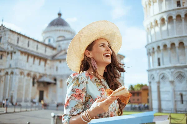 smiling young woman in floral dress with pizza and hat near Leaning Tower in Pisa, Italy.