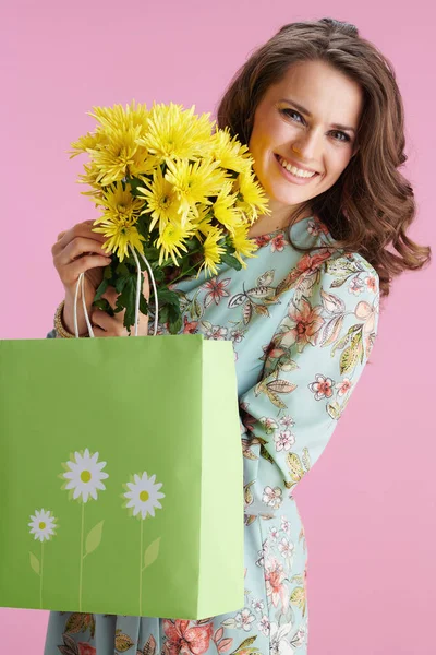 幸せなトレンディーな中年女性とともに長い波ブルネット髪とともに黄色菊の花と緑のショッピングバッグに対してピンクの背景 — ストック写真