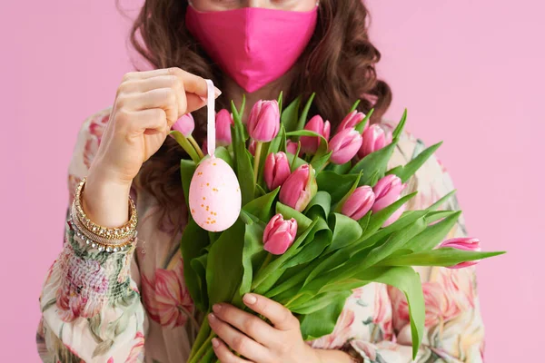 Closeup on middle aged woman with tulips bouquet, pink mask and easter egg isolated on pink background.