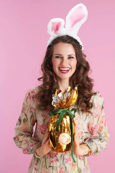 若い女性です花のドレスでウサギの耳とピンクの背景に隔離された黄金のイースターエッグ — ストック写真