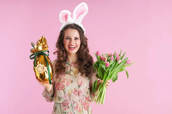 中年女性で花のドレスにチューリップ花束 ウサギの耳とピンクの背景に隔離された黄金のイースターエッグ — ストック写真
