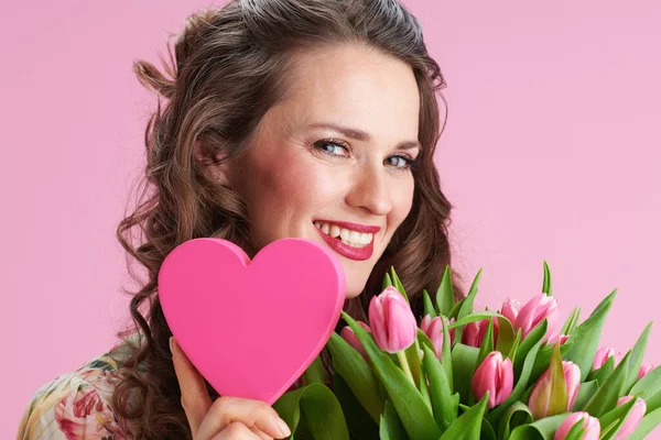 肖像画の笑顔エレガントな中年女性で花のドレスでチューリップ花束とピンクの心に対してピンクの背景 — ストック写真