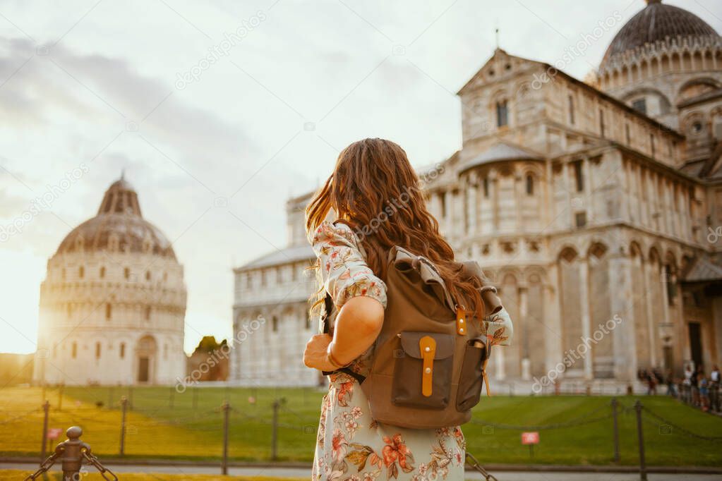 Seen from behind modern traveller woman in floral dress with backpack having excursion near Pisa Cathedral.