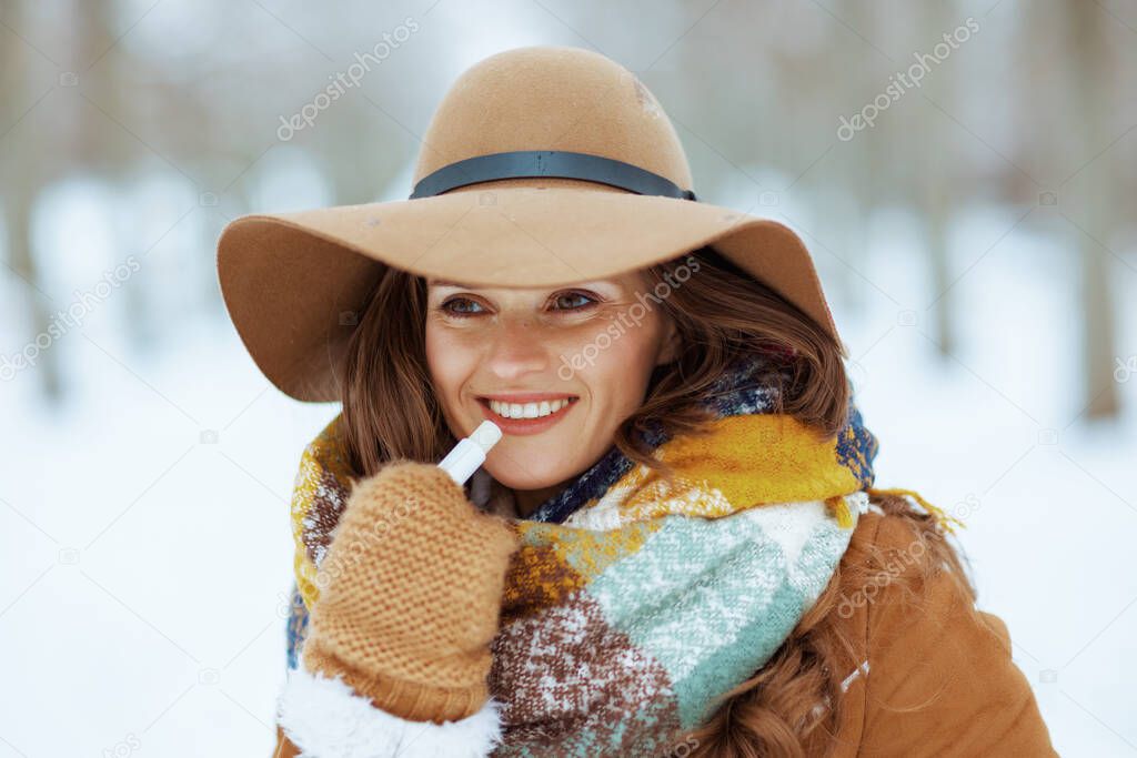 happy stylish woman in brown hat and scarf with mittens and hygienic lipstick in sheepskin coat outside in the city park in winter.