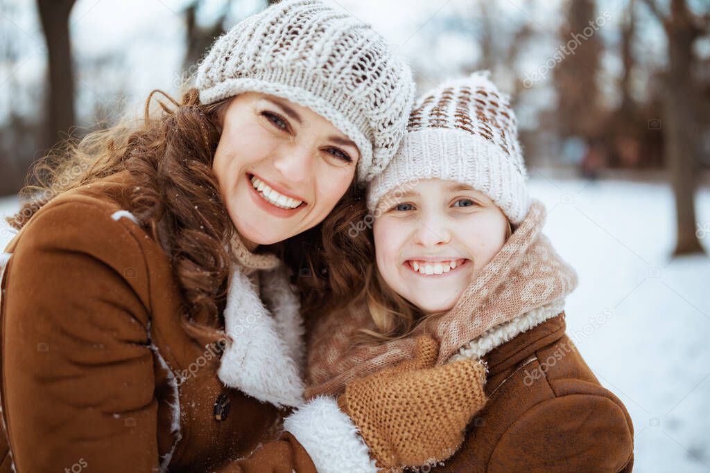 Portrait of happy stylish mother and child in a knitted hats and sheepskin coats with mittens in a knitted hat and sheepskin coat outdoors in the city park in winter.