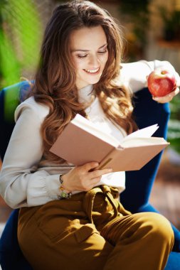 Yeşil Ev. Mutlu, orta yaşlı, uzun dalgalı saçlı, yeşil pantolonlu kırmızı elmalı ve güneşli bir günde modern evde gri bluzlu bir kadın..