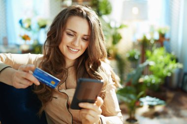 Yeşil Ev. Mutlu, modaya uygun, orta yaşlı, uzun dalgalı saçlı, kredi kartlı bir kadın. Güneşli bir günde modern evdeki e-ticaret sitesinde online alışveriş yapıyor..