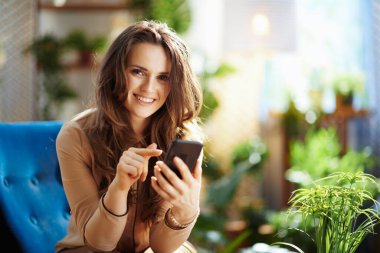 Yeşil Ev. Güneşli bir günde modern oturma odasında uzun dalgalı saçlı, modaya uygun bir ev hanımı akıllı telefon kullanarak mesaj gönderiyor..