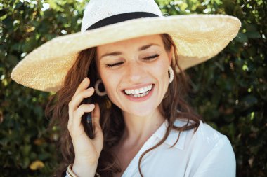 Beyaz gömlekli ve şapkalı mutlu orta yaşlı ev hanımı açık havada, yeşil duvarın yanında bir akıllı telefondan konuşuyor..