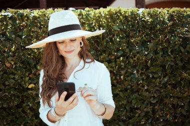 Yeşil duvarın yanında akıllı telefon uygulamaları kullanarak beyaz tişörtlü, gülümseyen modern kadın..