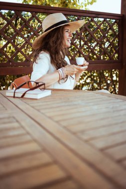 Beyaz gömlekli, kitap, şapka ve gözlüklü 40 yaşında bir kadın otel odasının terasında kahve içerken gülümsüyor..