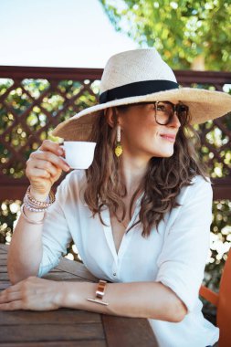 Beyaz tişörtlü, kahve, şapka ve gözlüklü, modaya uygun orta yaşlı kadın otel odasının avlusunda oturuyor..