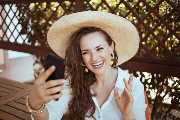 幸せな現代40歳の女性の白いシャツを着て帽子を持っていますウェブカメラミーティングゲストハウスホテルのテラスでスマートフォン — ストック写真