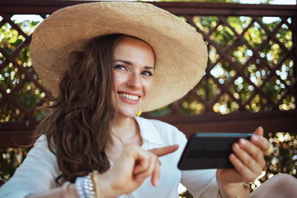 パティオでスマートフォンを使ってテキストメッセージを送る帽子付きの白いシャツを着た中高年主婦 — ストック写真