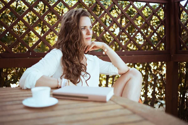 身穿白衬衫 头戴咖啡杯 头戴书本的温文尔雅女人坐在阳台的桌子旁 — 图库照片