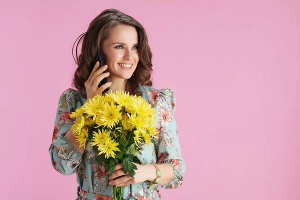 幸せな流行の女性で花のドレスとともに黄色菊の花ピンクの背景に隔離されたスマートフォンで話している — ストック写真