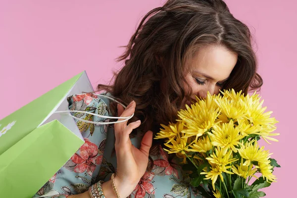 幸せな40歳の女性とともに長い波状ブルネット髪とともに黄色菊の花と緑のショッピングバッグに対してピンクの背景 — ストック写真