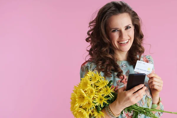 黄色の菊の花とクレジットカードで花のドレスでスタイリッシュな女性の笑顔の肖像ピンクの背景にスマートフォンを使用してテキストメッセージを送信 — ストック写真