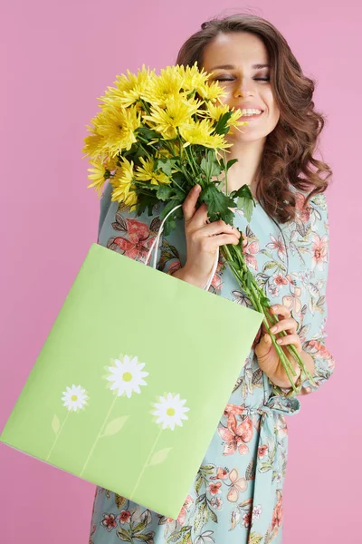 幸せな若いです若いです女性とともに長い波状ブルネット髪とともに黄色菊の花と緑のショッピングバッグ隔離されたピンクの背景 — ストック写真