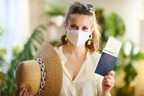 コロナウイルスのパンデミックの間の旅行 晴れた日に現代の家の中で鮮やかなパスポート 医療マスク 航空券 麦藁帽子とサングラスを持つスタイリッシュなソロ観光女性 — ストック写真