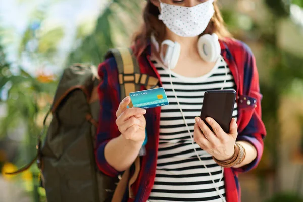 Travel during covid-19 pandemic. Closeup on young tourist woman with white medical mask, backpack, credit card and smartphone booking trip online in the modern living room in sunny day.
