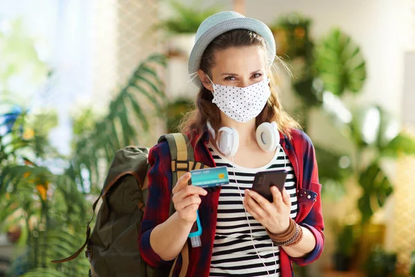 コロナウイルスのパンデミックの間の旅行 白い医療マスク バックパック ヘッドフォン クレジットカードやスマートフォンの予約旅行でオンラインで現代のリビングルームで晴れた日に — ストック写真