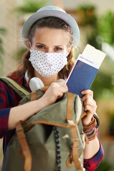 Travel during coronavirus pandemic. Portrait of modern woman with covid passport, white medical mask, backpack and hat in the modern living room in sunny day.
