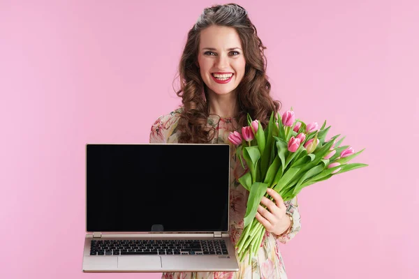 現代の女性を笑顔です花のドレスにチューリップ花束ラップトップの空の画面を示すピンクの背景 — ストック写真