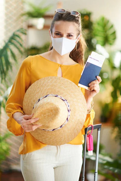 Travel during coronavirus pandemic. modern tourist woman with covid passport, medical mask, air ticket, wheel bag, sanitiser and straw hat at modern home in sunny day.