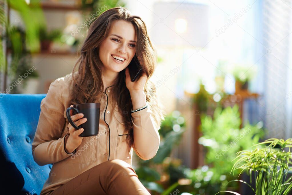 Green Home. happy 40 years old woman with long wavy hair with cup of hot beverage using a smartphone in the modern living room in sunny day.