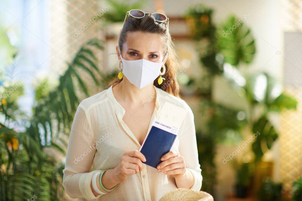 Travel during coronavirus pandemic. trendy tourist woman with covid passport, medical mask, air ticket and sunglasses at modern home in sunny day.