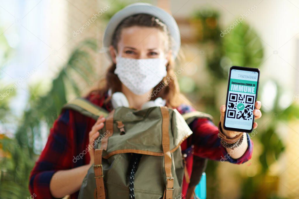 Travel during covid-19 pandemic. Closeup on trendy solo tourist woman with smartphone, covid passport app, white medical mask and backpack in the modern house in sunny day.