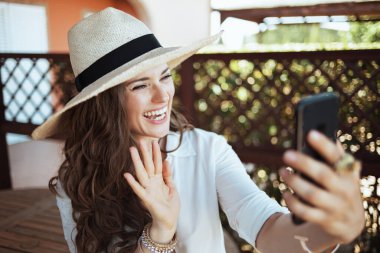 Beyaz tişörtlü mutlu bir bayan otel odasının terasındaki akıllı telefondan video görüşmesi yapıyor..