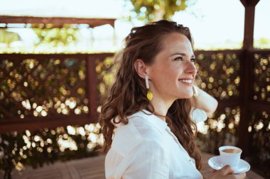 Kırk yaşındaki beyaz gömlekli ev hanımı verandada kahve içiyor..